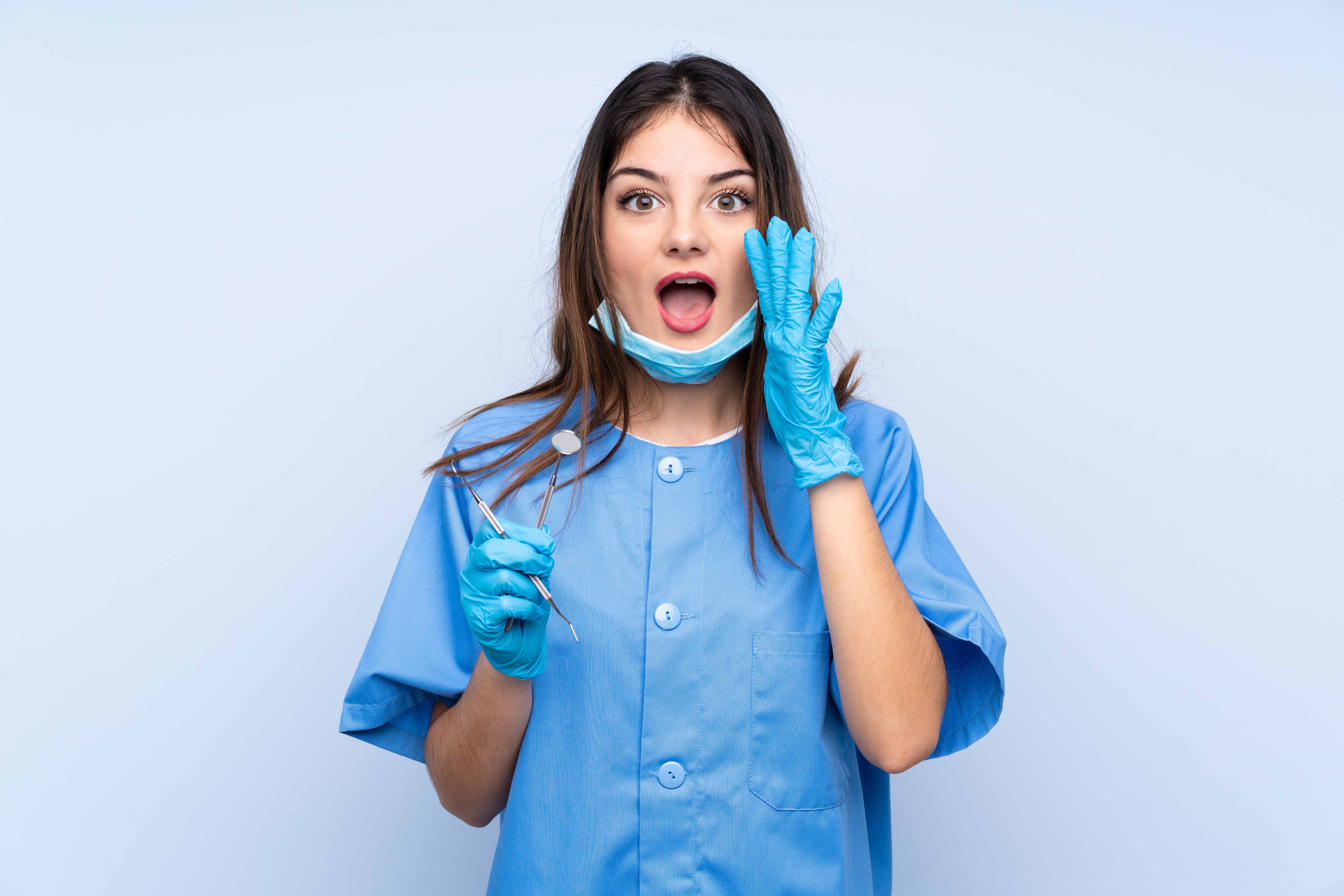 水色の白衣・手袋をした女性歯科助手が、右手に手に歯の治療器具を持ち、左手を口元に当てて呼びかけている。